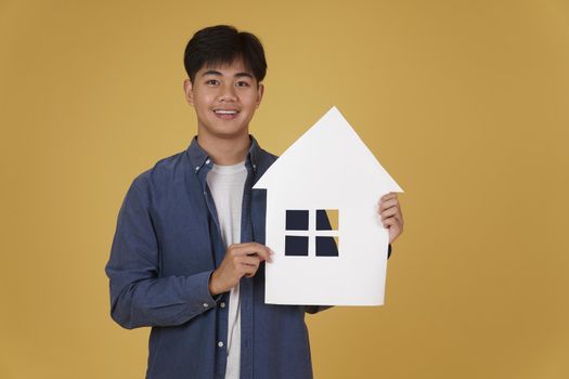 portrait of smiling happy cheerful young asian man dressed casually with home house paper cutout isolated on yellow studio background. real estate purchase concept