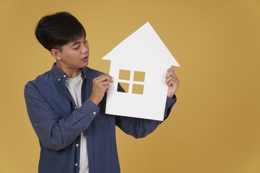 portrait of smiling happy cheerful young asian man dressed casually with home house paper cutout isolated on yellow studio background. real estate purchase concept