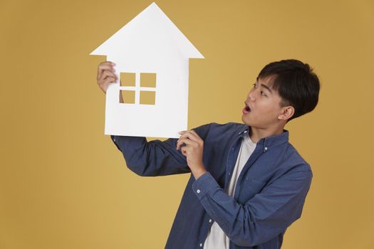 portrait of smiling happy cheerful young asian man dressed casually with home house paper cutout isolated on yellow studio background. real estate purchase concept