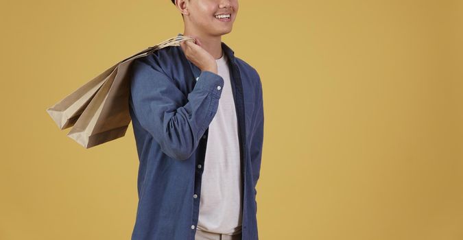 portrait of happy young asian man dressed casually holding shopping bags isolated on yellow studio background