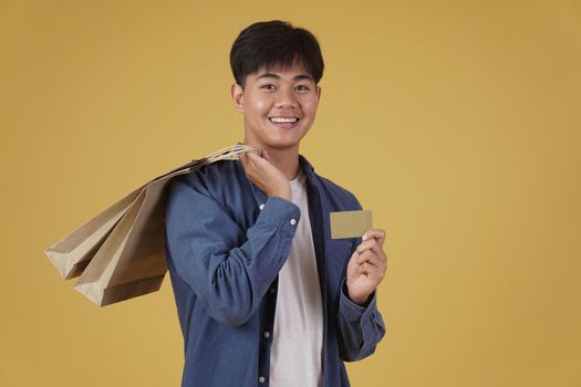 portrait of happy young asian man dressed casually holding shopping bags and credit card isolated on yellow studio background