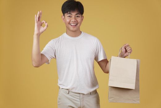 portrait of happy young asian man dressed casually holding shopping bags isolated on yellow studio background