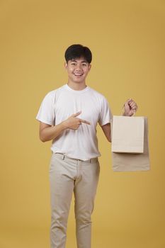 portrait of happy young asian man dressed casually holding shopping bags isolated on yellow studio background