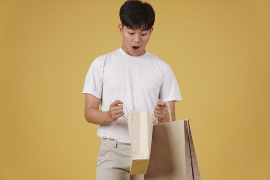 portrait of surprised excited young asian man dressed casually opening shopping bags isolated on yellow studio background
