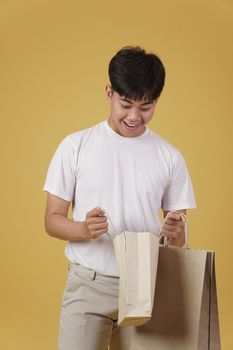 portrait of happy young asian man dressed casually opening shopping bags isolated on yellow studio background