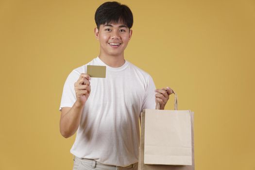 portrait of happy young asian man dressed casually holding shopping bags and credit card isolated on yellow studio background