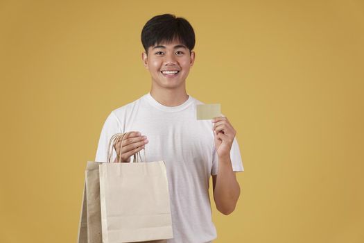 portrait of happy young asian man dressed casually holding shopping bags and credit card isolated on yellow studio background