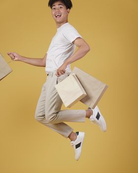 portrait of happy excited young asian man dressed casually holding shopping bags jumping isolated on yellow studio background