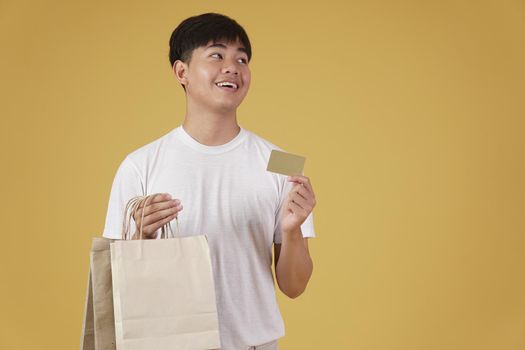 portrait of happy young asian man dressed casually holding shopping bags and credit card isolated on yellow studio background