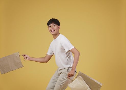 portrait of happy young asian man dressed casually holding shopping bags isolated on yellow studio background