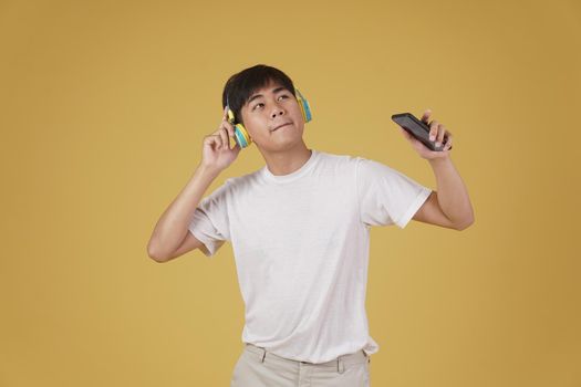 portrait of happy joyful cheerful young asian man wearing headphones listening to music on smartphone and dancing isolated on yellow studio background