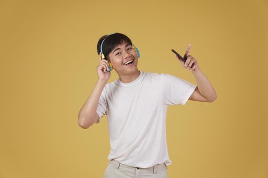 portrait of happy joyful cheerful young asian man wearing headphones listening to music on smartphone and dancing isolated on yellow studio background