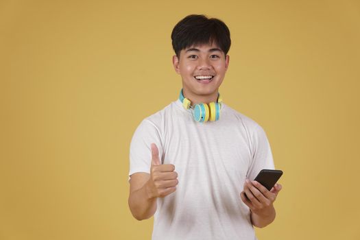 portrait of happy joyful cheerful young asian man wearing headphones listening to music on smartphone and dancing isolated on yellow studio background