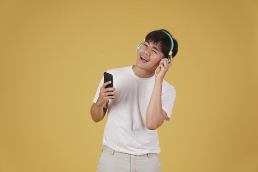 portrait of happy joyful cheerful young asian man wearing headphones listening to music on smartphone and dancing isolated on yellow studio background