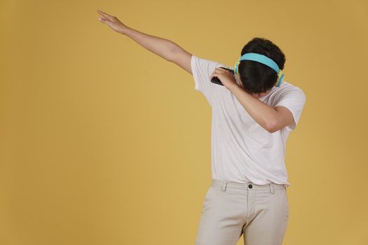 portrait of happy joyful cheerful young asian man wearing headphones listening to music on smartphone and dancing isolated on yellow studio background