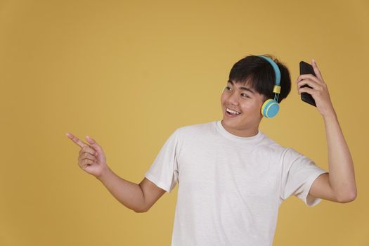 portrait of happy joyful cheerful young asian man wearing headphones listening to music on smartphone and dancing isolated on yellow studio background