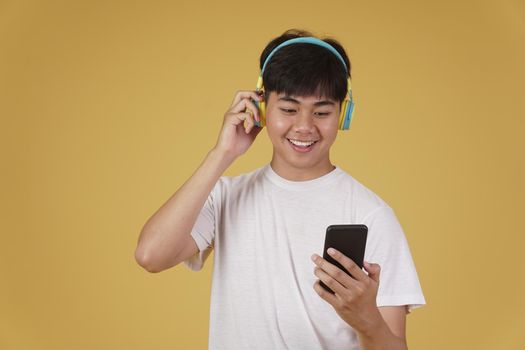 portrait of happy joyful cheerful young asian man wearing headphones listening to music on smartphone and dancing isolated on yellow studio background