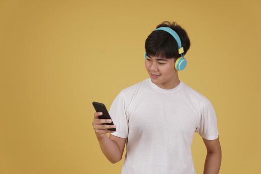 portrait of happy joyful cheerful young asian man wearing headphones listening to music on smartphone and dancing isolated on yellow studio background