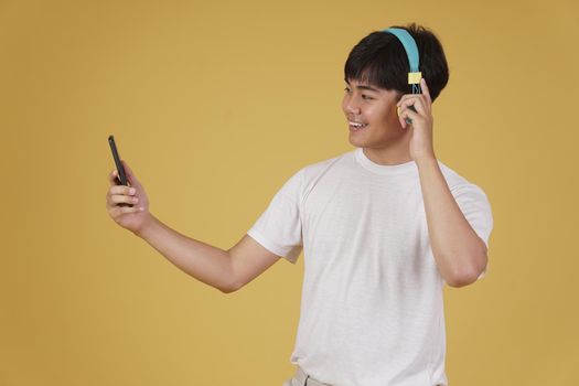 portrait of happy joyful cheerful young asian man wearing headphones listening to music on smartphone and dancing isolated on yellow studio background