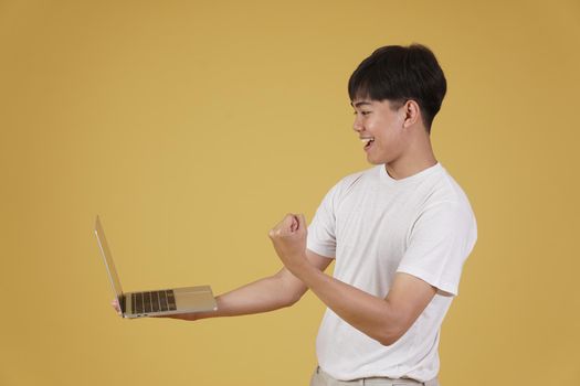 portrait of happy glad rejoicing cheerful young asian man dressed casually holding laptop computer doing fist winner gesture isolated on yellow studio background