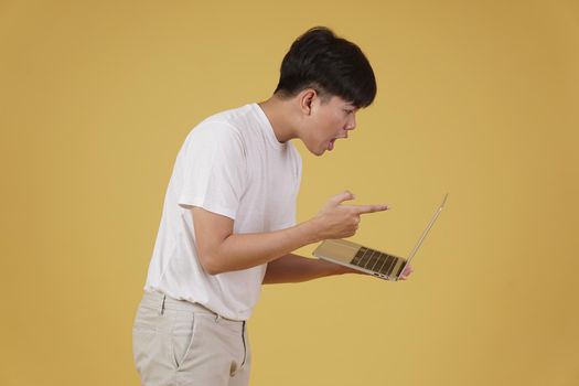 aggressive angry upset furious young asian man scolding screaming yelling on laptop computer isolated on yellow studio background
