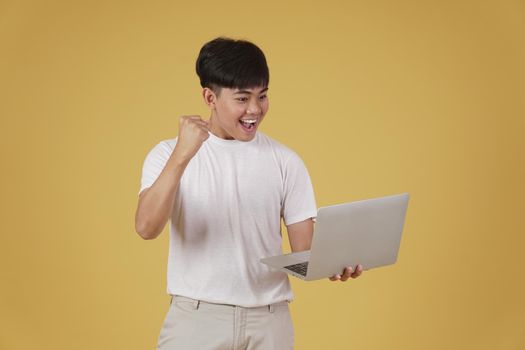 portrait of happy glad rejoicing cheerful young asian man dressed casually holding laptop computer doing fist winner gesture isolated on yellow studio background