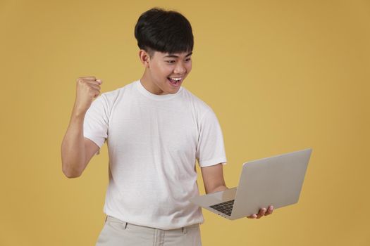 portrait of happy glad rejoicing cheerful young asian man dressed casually holding laptop computer doing fist winner gesture isolated on yellow studio background