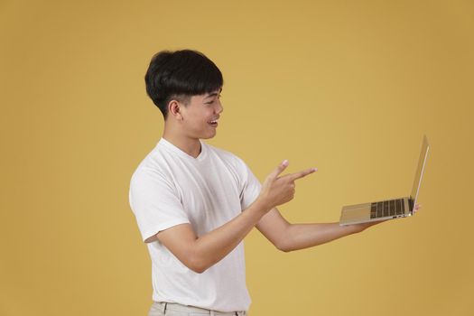 happy glad rejoicing cheerful young asian man dressed casually holding pointing finger at laptop computer screen isolated on yellow studio background