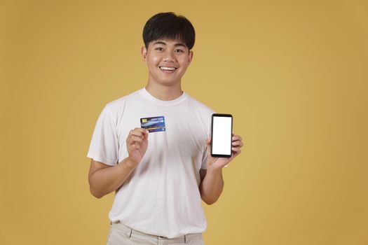 portrait of happy young asian man dressed casually holding smartphone and credit card for shopping online isolated on yellow studio background