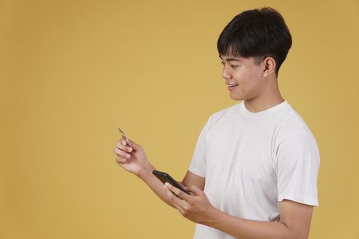 portrait of happy young asian man dressed casually holding smartphone and credit card for shopping online isolated on yellow studio background