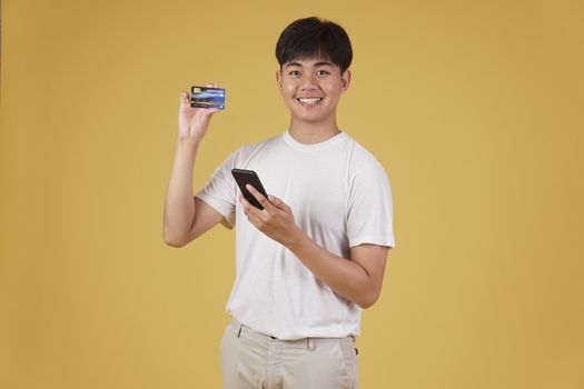 portrait of happy young asian man dressed casually holding smartphone and credit card for shopping online isolated on yellow studio background