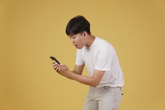 portrait of shocked surprised excited young asian man dressed casually looking at mobile smart phone isolated on yellow studio background