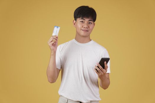 portrait of happy young asian man dressed casually holding smartphone and credit card for shopping online isolated on yellow studio background