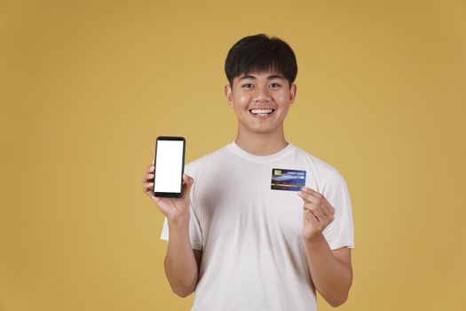 portrait of happy young asian man dressed casually holding smartphone and credit card for shopping online isolated on yellow studio background