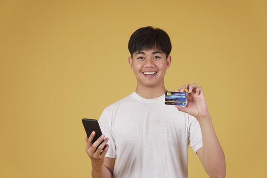 portrait of happy young asian man dressed casually holding smartphone and credit card for shopping online isolated on yellow studio background