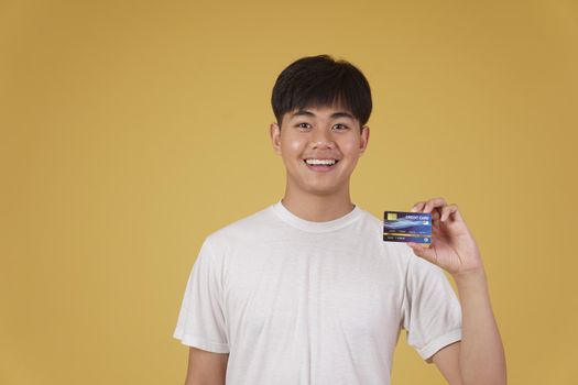 portrait of happy young asian man dressed casually holding credit card for shopping payment isolated on yellow studio background