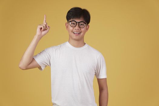 happy cheerful young asian man student dressed casually wearing eyeglasses pointing finger up having idea isolated on yellow studio background