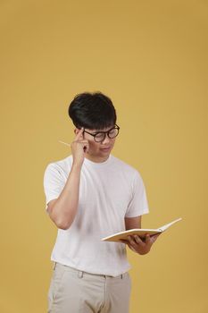 thoughtful young asian man student dressed casually wearing eyeglasses holding notebook thinking isolated on yellow studio background