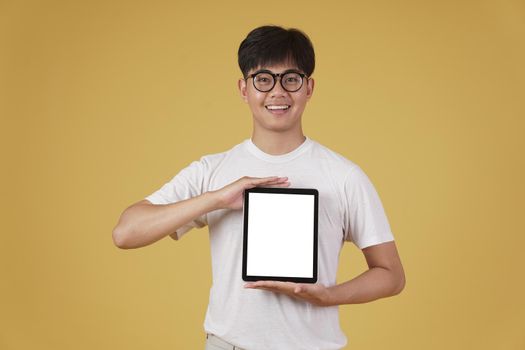 portrait of smiling happy cheerful young asian man dressed casually showing blank digital tablet screen isolated on yellow studio background.