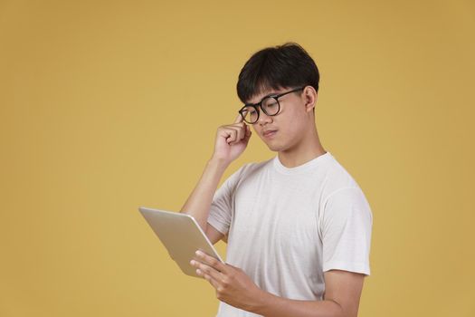 thoughtful young asian man dressed casually thinking using digital tablet isolated on yellow studio background.