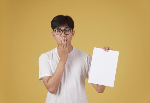 portrait of surprised young asian man dressed casually showing blank empty placard paper isolated on yellow studio background.