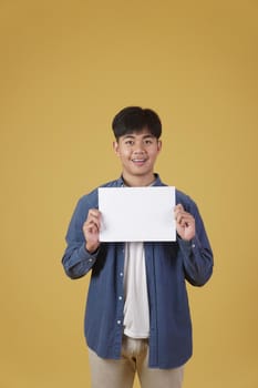 portrait of smiling happy cheerful young asian man dressed casually showing blank empty placard paper isolated on yellow studio background.