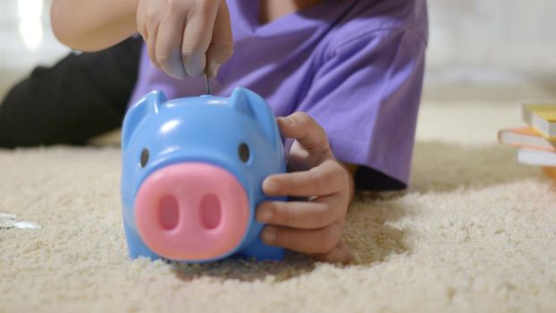 Happy Asian kid boy preschool putting pin money coins into blue faced piglet slot. Little child putting coin into piggy bank for saving with pile of coins at home, Investment education concept