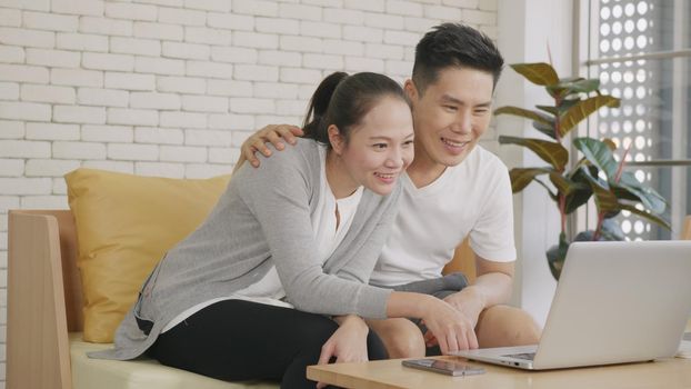 Happy Asian family couple husband and wife laughing sitting on sofa using laptop computer webcam technology talking making online social distance video call to their children at home.