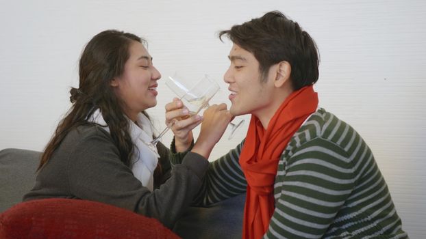 Happy Asian beautiful young family couple husband and wife on sofa in home living room, man and woman sit sofa in love clink glasses with champagne enjoy and drink winter holidays together