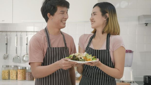 Happy Asian beautiful family couple husband and wife cooking vegetable salad in kitchen together at home. The man and woman handing a plate of salad to the camera. healthy food concept,
