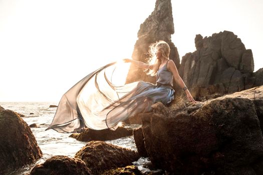 Beautiful European girl in a long dress sits on the rocks by the sea