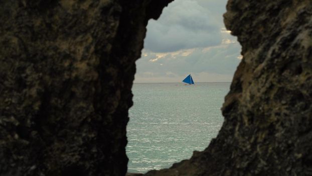 Sailing boat view through a hole in the rock. Sailing ship yachts with blue sails in the ocean. Sail boat on sea. Philippines, Boracay. Travel concept.