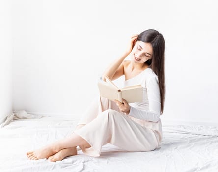 Beautiful woman in white cozy clothes reading a book sitting on the floor