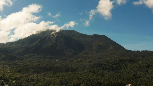 Mountains covered rainforest, trees and blue sky with clouds, aerial view. Camiguin, Philippines. Mountain landscape on tropical island with mountain peaks covered with forest. Slopes of mountains with evergreen vegetation.